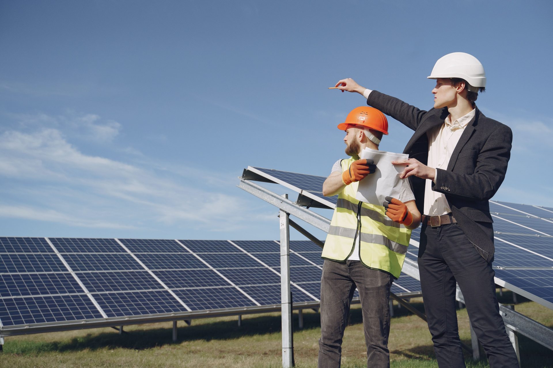 Junger Geschäftsmann in formeller Kleidung und Polier in gelber Weste blicken vor Solarmodulen zur Seite, während sie den Arbeitsablauf besprechen.