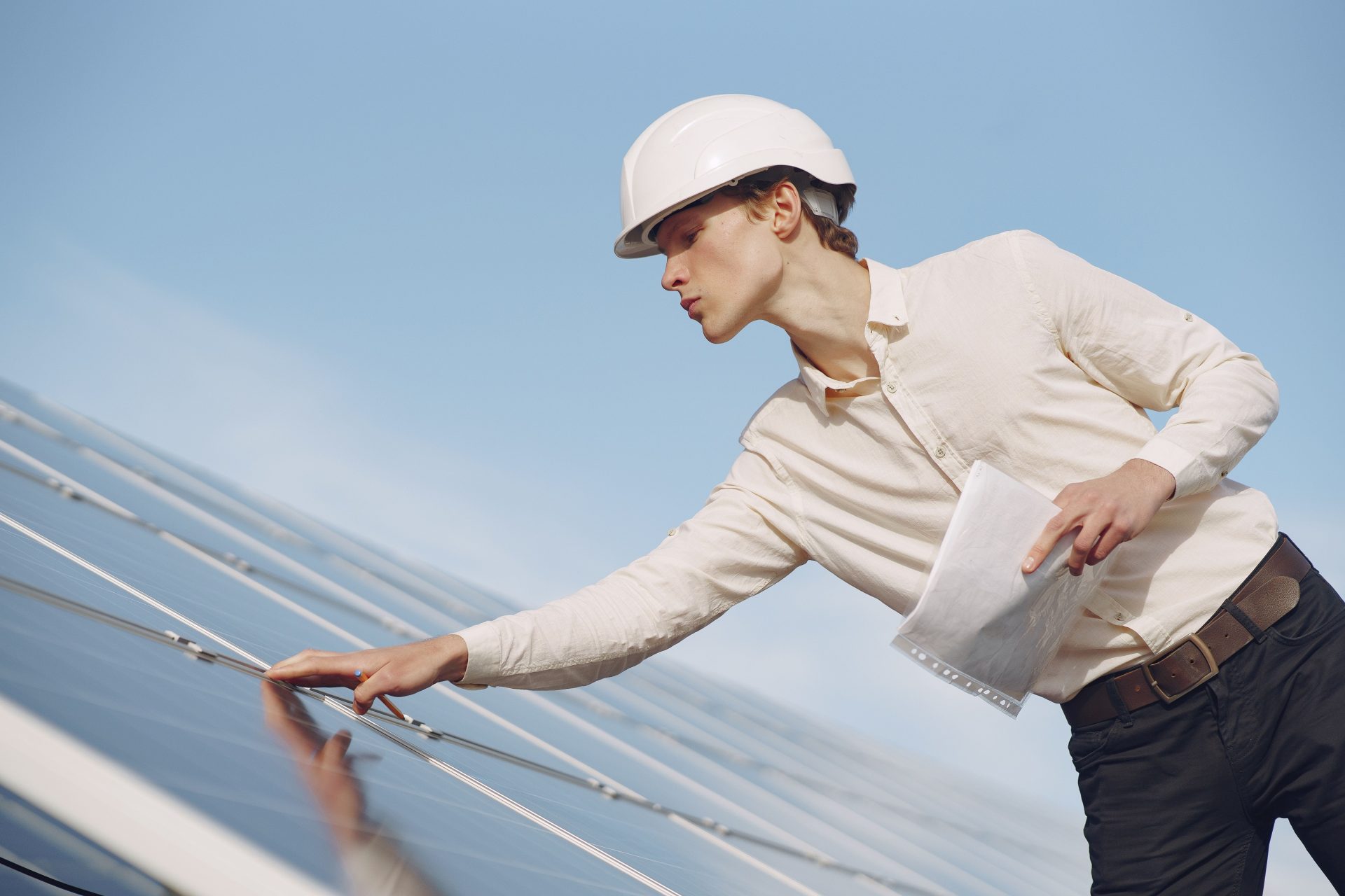 Ansicht aus der Froschperspektive: ein junger männlicher Inspektor in formeller Kleidung und mit Schutzhelm steht vor Solarmodulen und überprüft Details der Konstruktion.