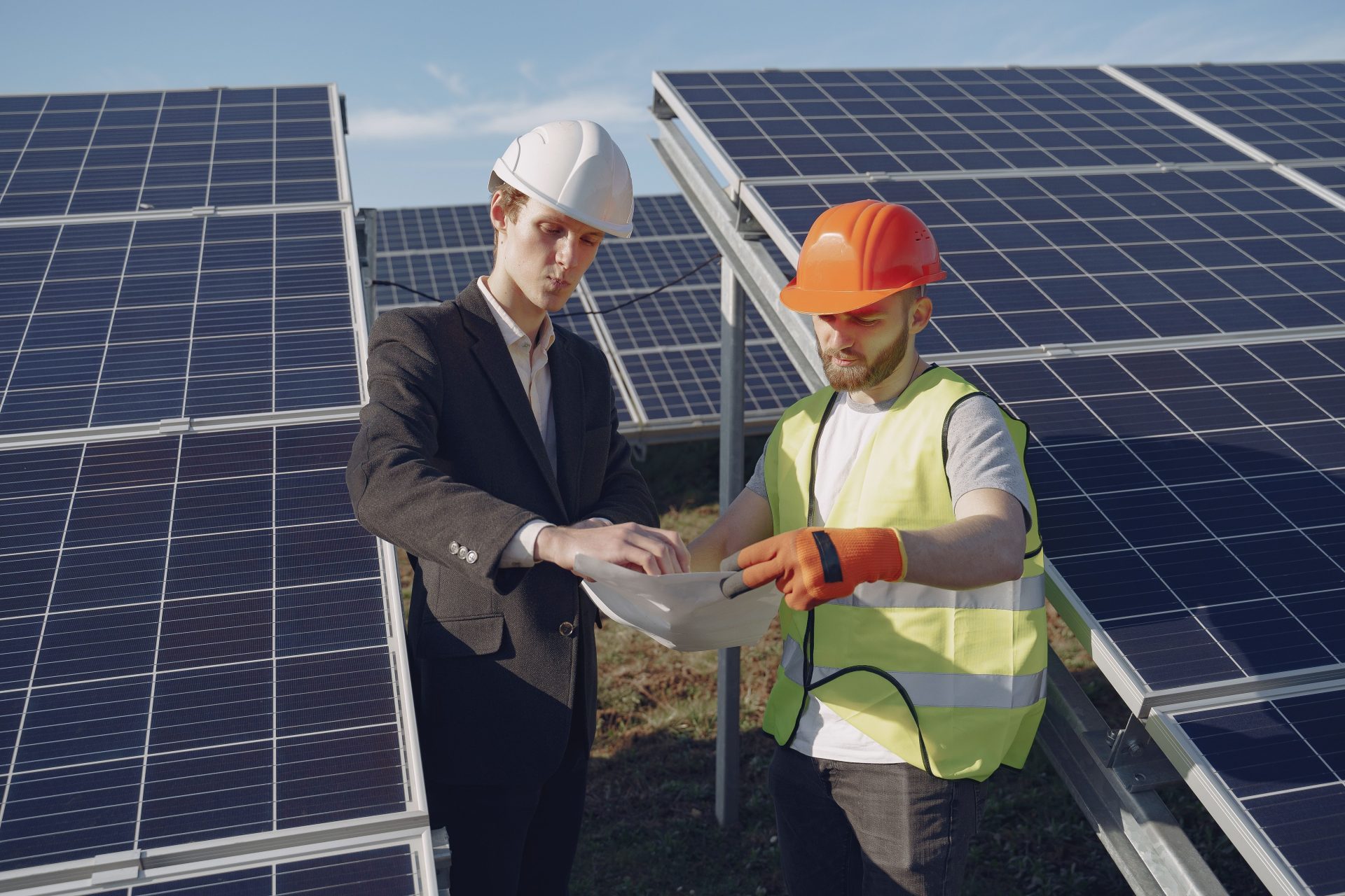 Konzentrierter junger Geschäftsmann und Vorarbeiter mit Schutzhelmen stehen zwischen Solarmodulen und prüfen technische Unterlagen.