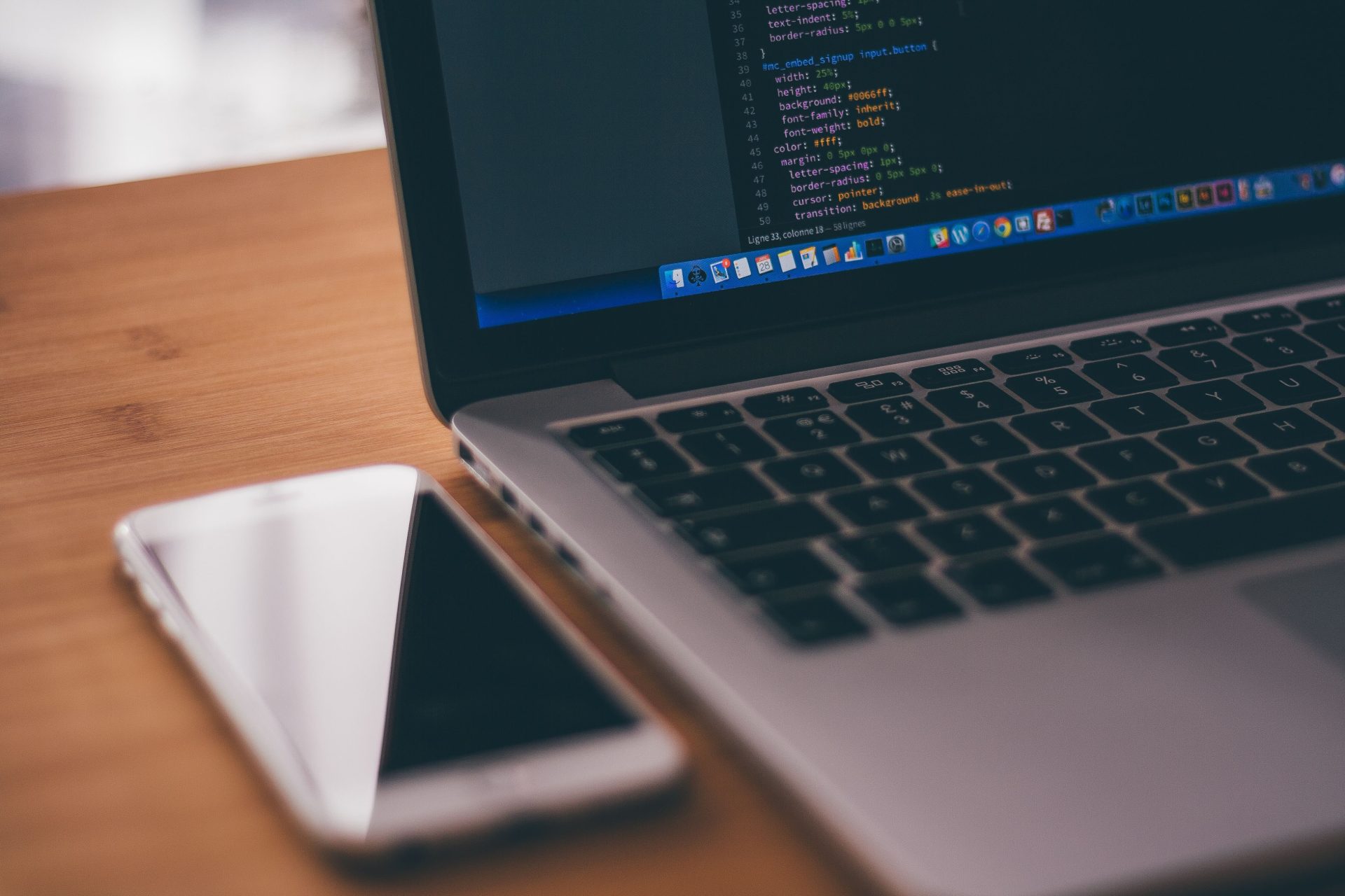 Programmier-Setup mit Laptop, der Code anzeigt, und Smartphone auf einem Holztisch – ideal für Themen aus dem Technologiebereich.